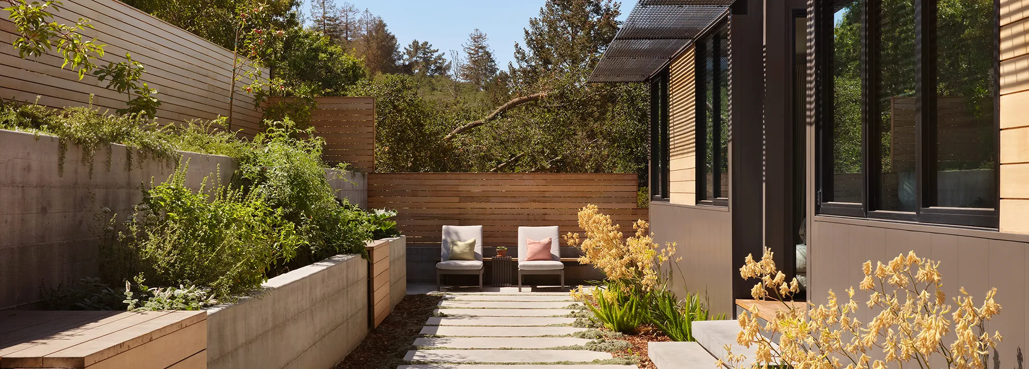 Modern backyard patio with two chairs, plants in raised concrete beds, and wooden privacy fence.