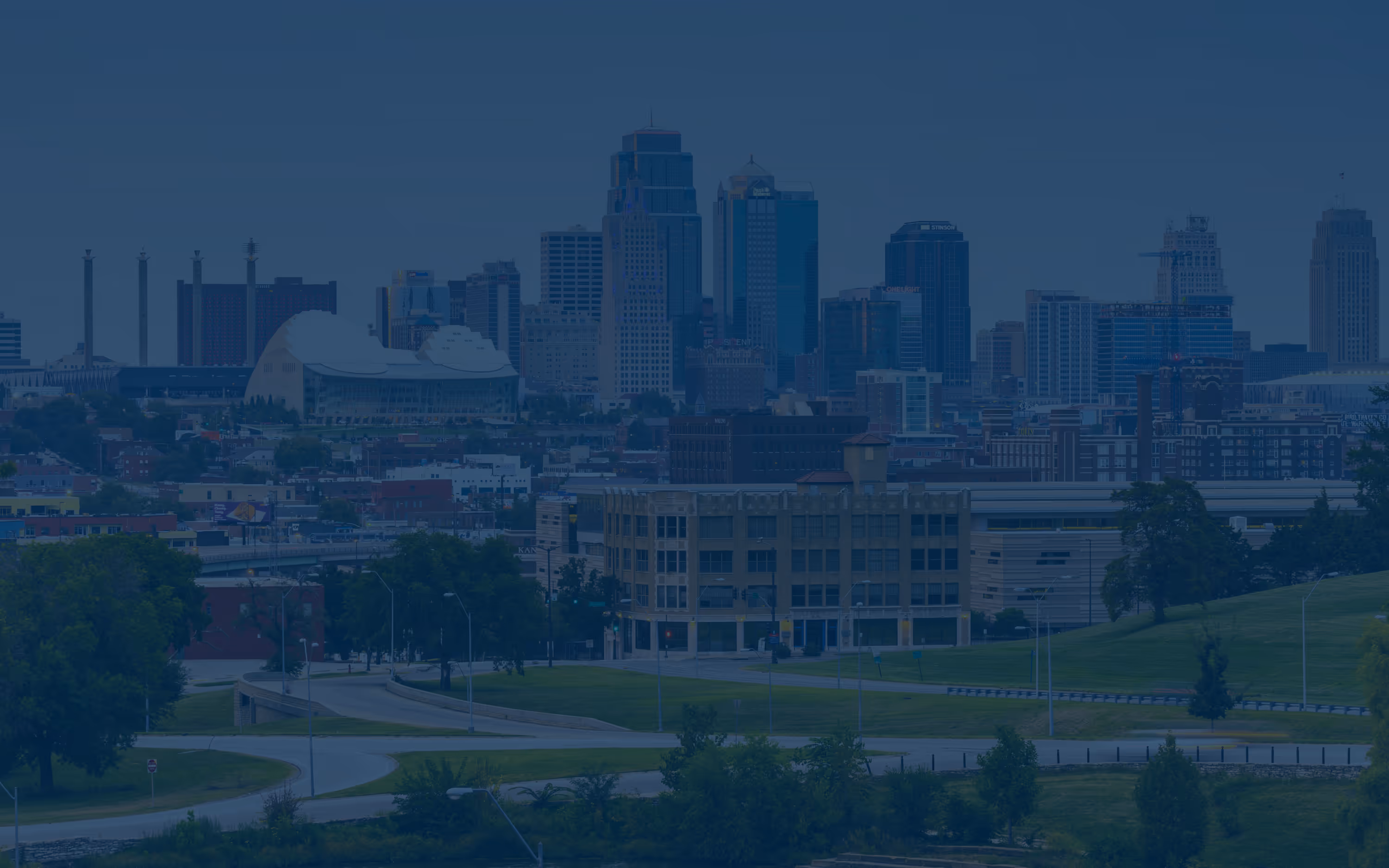 Cityscape of Kansas City with downtown skyscrapers, mid-rise buildings, and green parkland in the foreground.