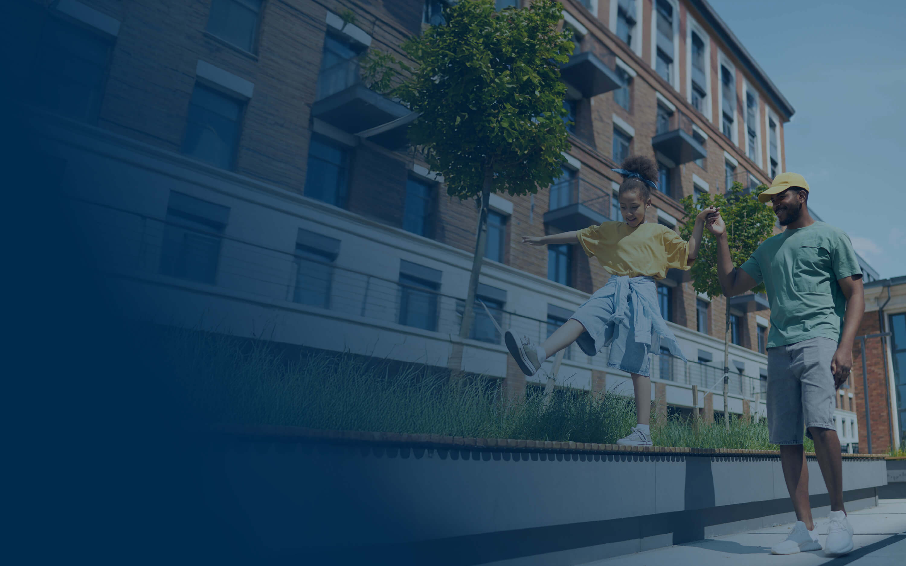 Man holding hands with a girl who is balancing on a low wall outside near a brick building and trees.