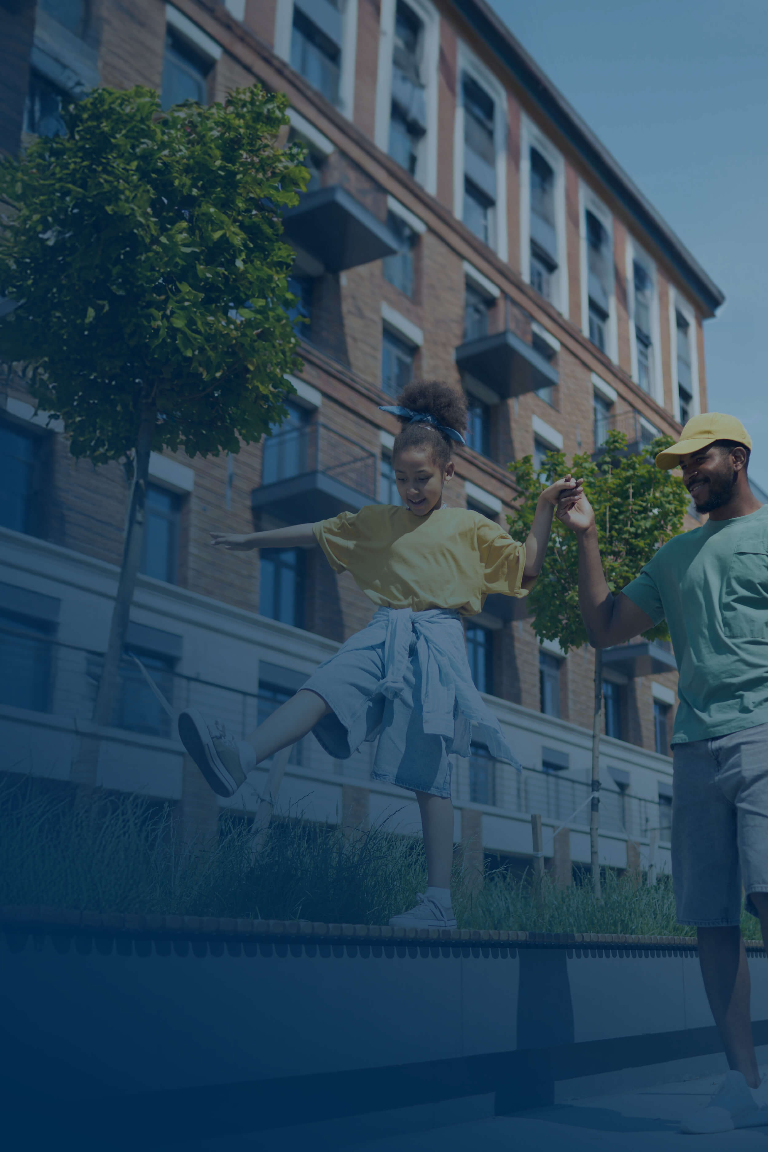 Man holding hands with a girl who is balancing on a low wall outside near a brick building and trees.