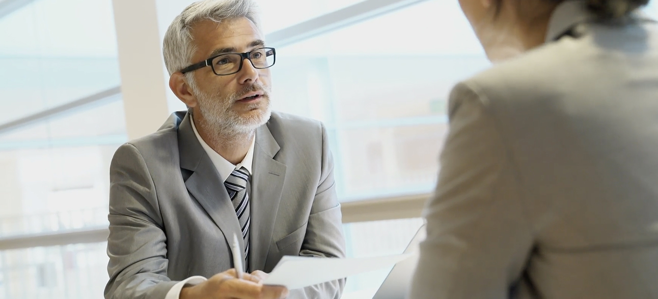 Mature businessman in gray suit and glasses holding a document, speaking to a woman in a modern office.