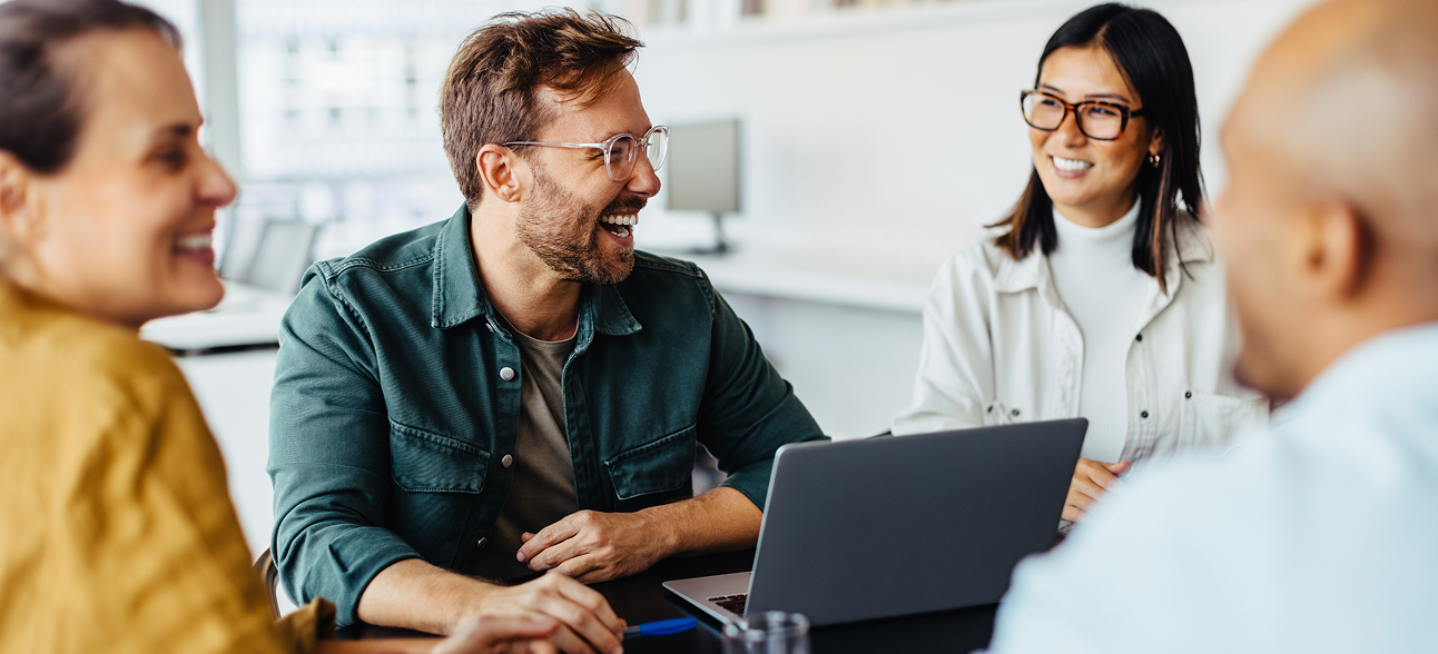 Four diverse colleagues smiling and talking around a laptop in a bright office.