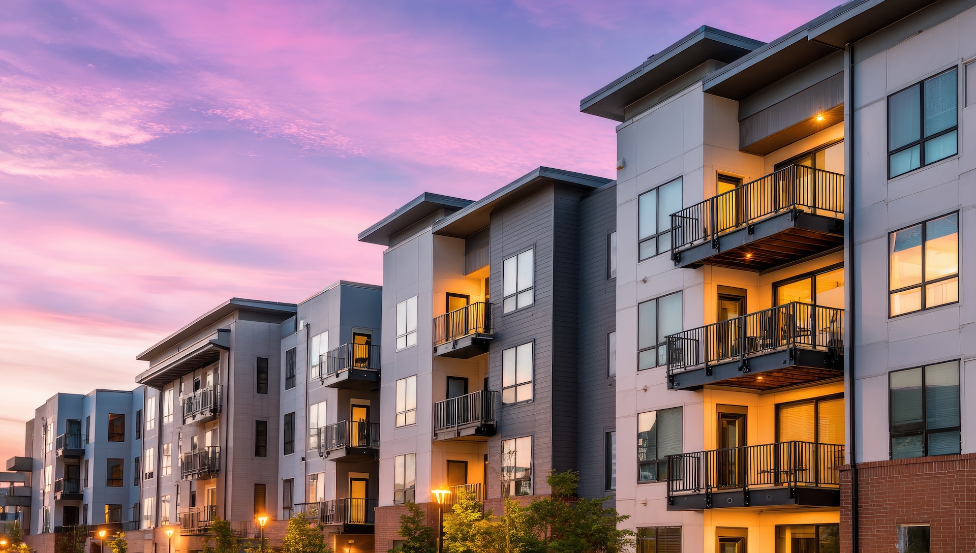 Modern apartment buildings with balconies lit by warm indoor lights at sunset under a purple and pink sky.