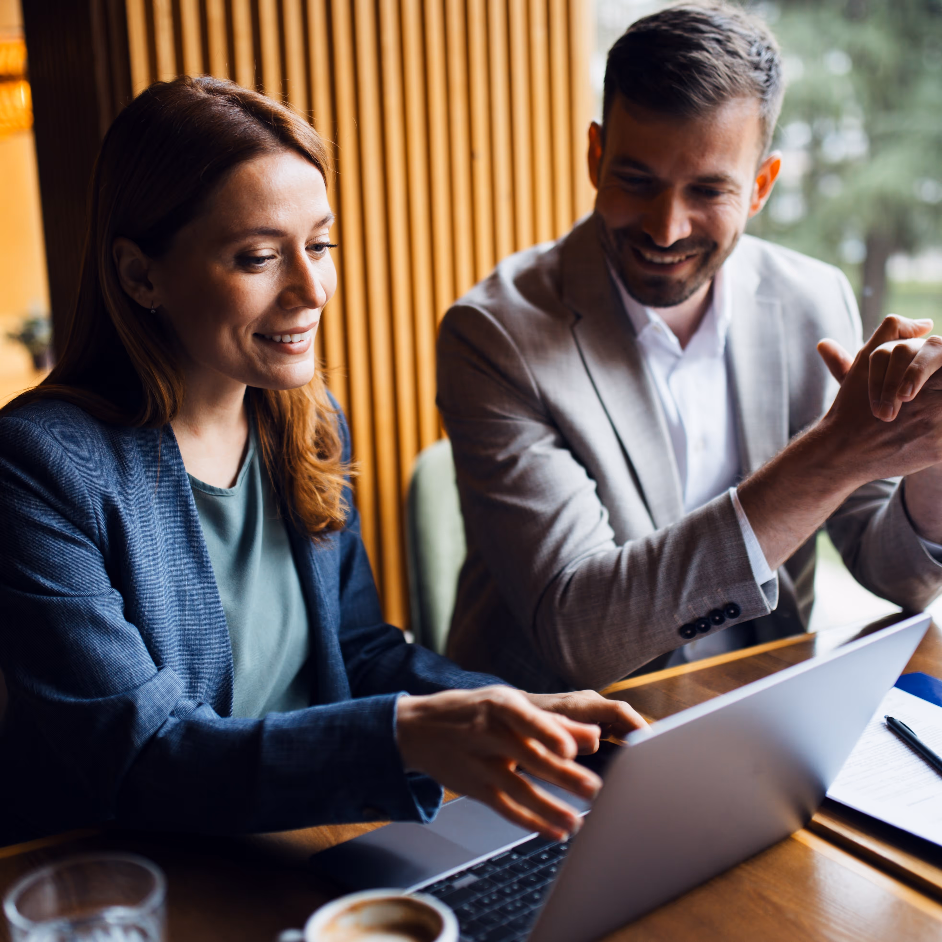 Two professionals in business attire smiling and reviewing information on a laptop at a wooden table.