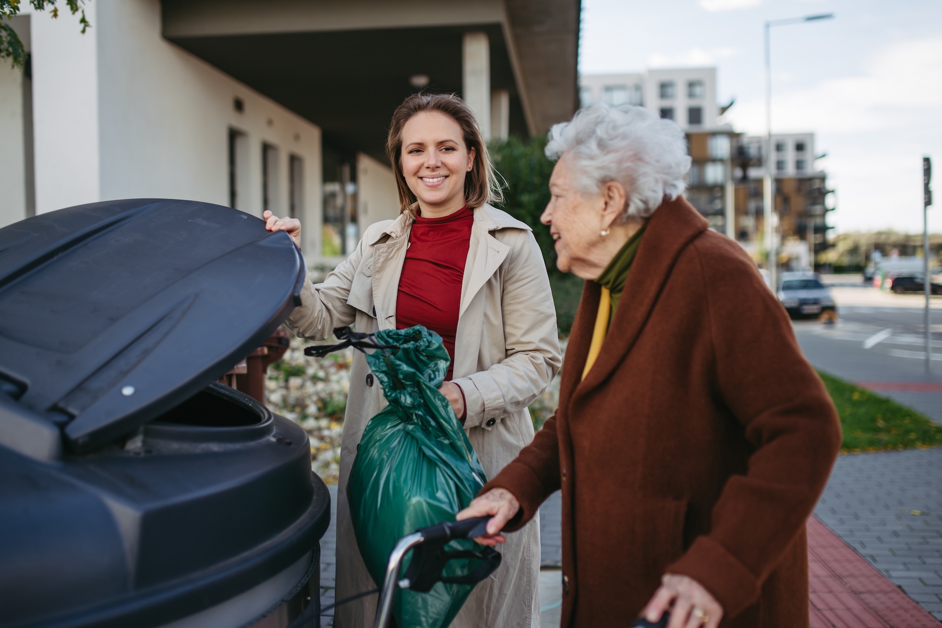 Young woman smiling while helping elderly woman dispose of a garbage bag in an outdoor trash bin.
