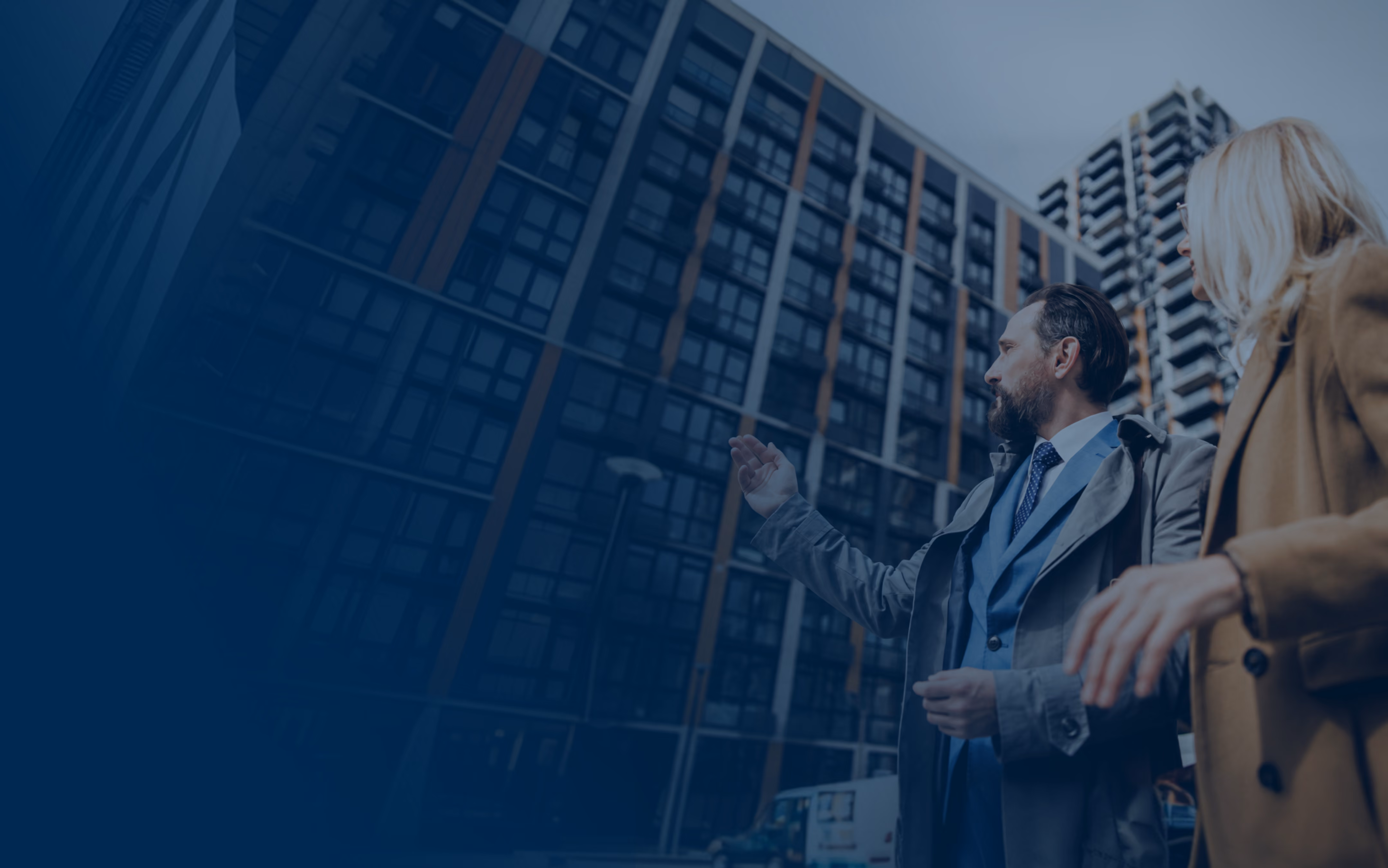 Two professionals, a man and a woman, stand outside in front of tall modern office buildings, with the man gesturing towards the buildings as if explaining something.