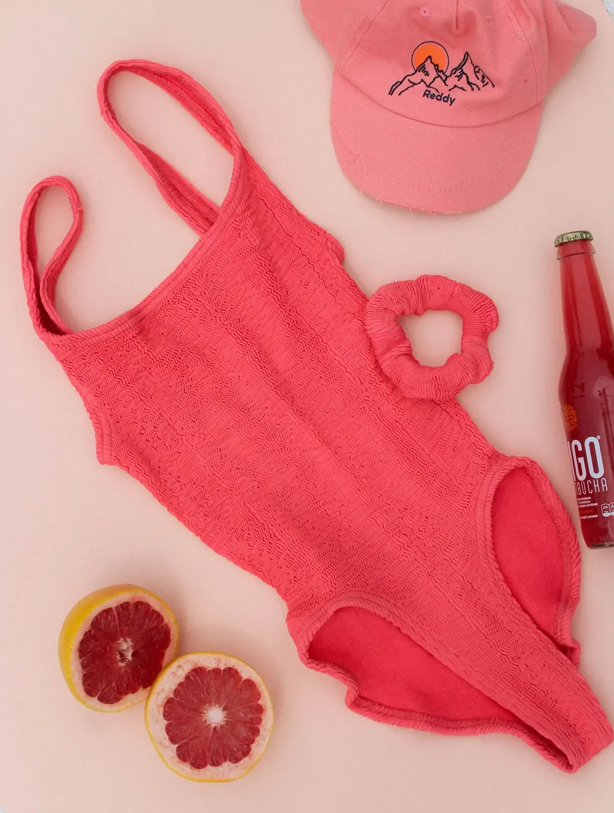 One piece pink swimsuit made out of Cienna lying flat, surrounded by fruit and accessories