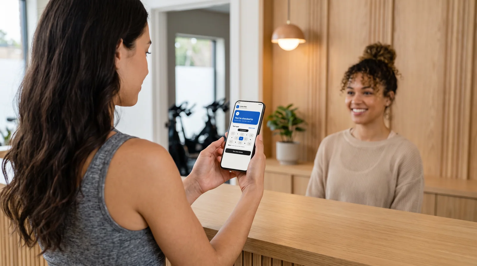 Member at a boutique studio front desk holding a phone showing her Walla check-in and Book a Spot bike 8, sample studio