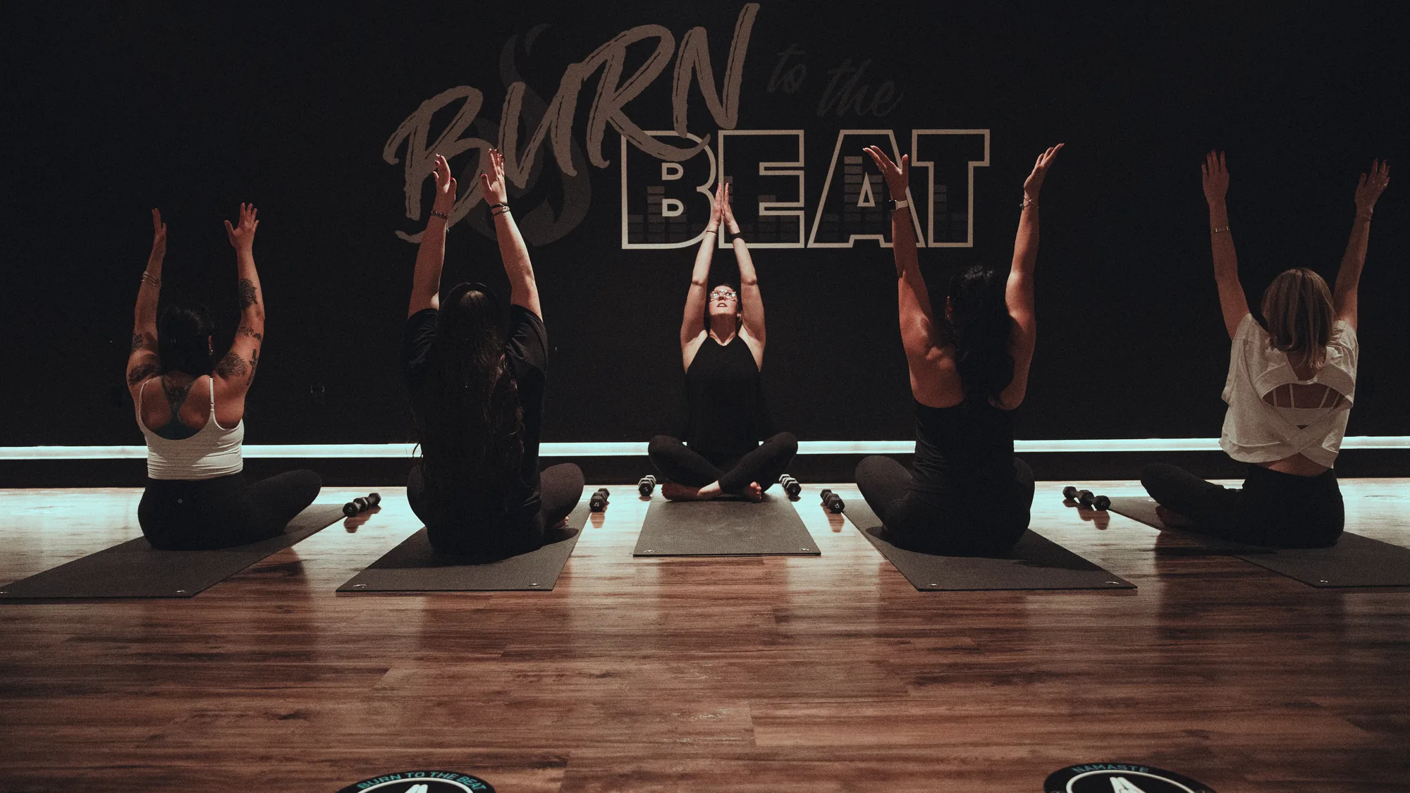 Instructor leading a seated mat class with arms raised in a dark, warm-lit boutique studio
