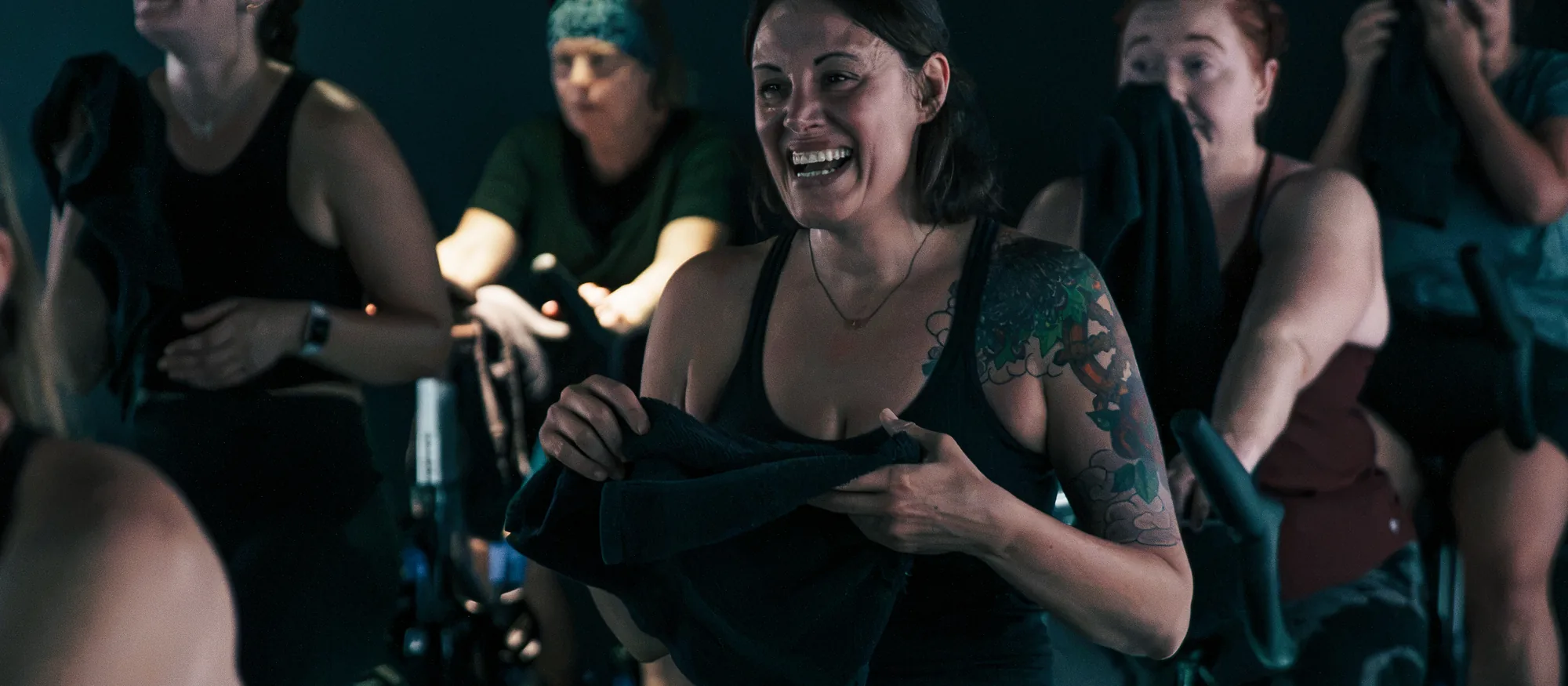 Riders laughing together during an indoor cycling class in a dim, teal-lit boutique studio