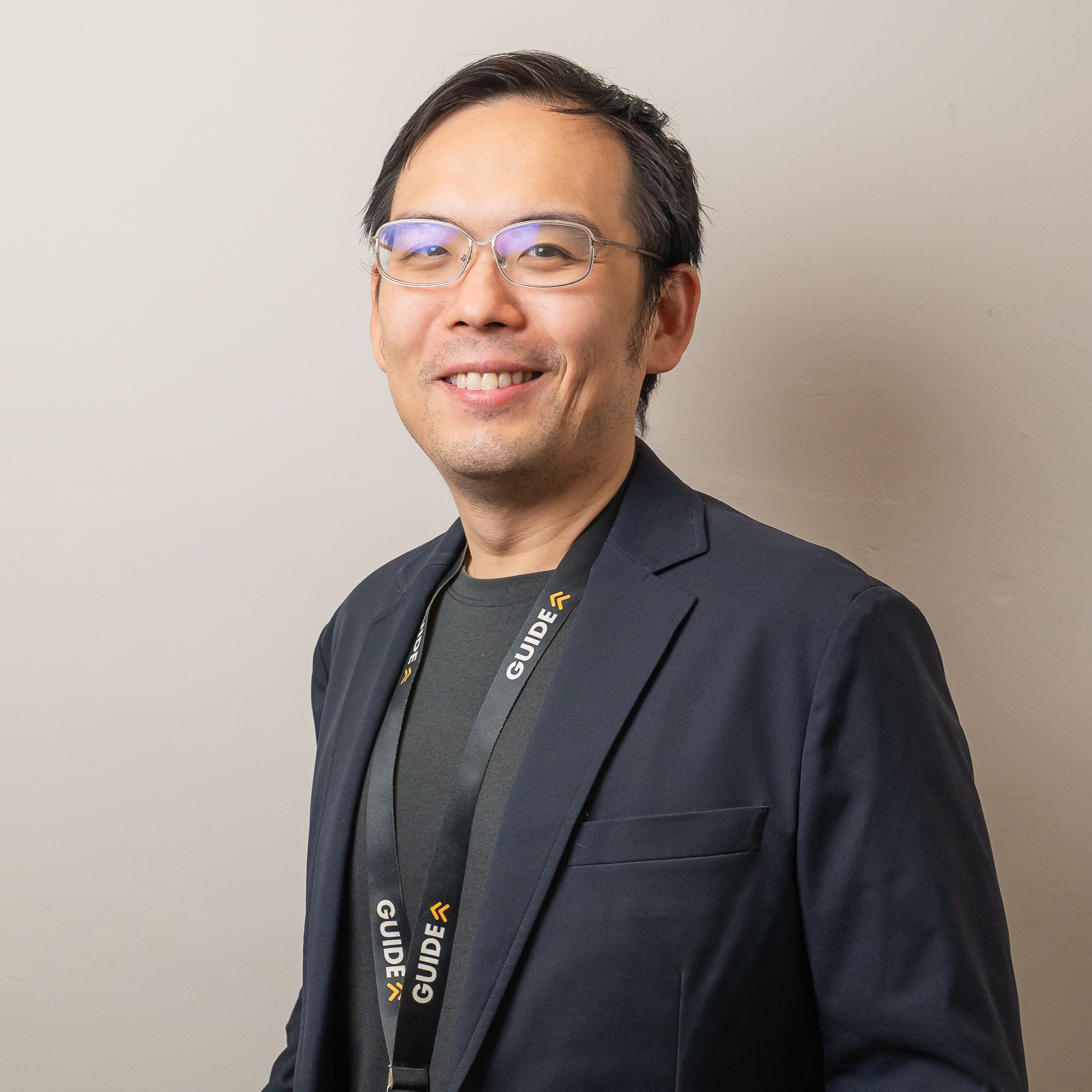 Man wearing glasses and a suit with a 'GUIDE' lanyard, standing indoors with hands clasped.