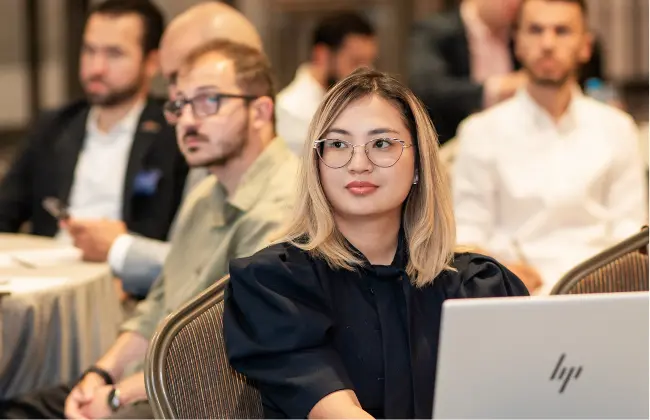 Woman with glasses sitting at a laptop in a meeting with other attendees in the background.