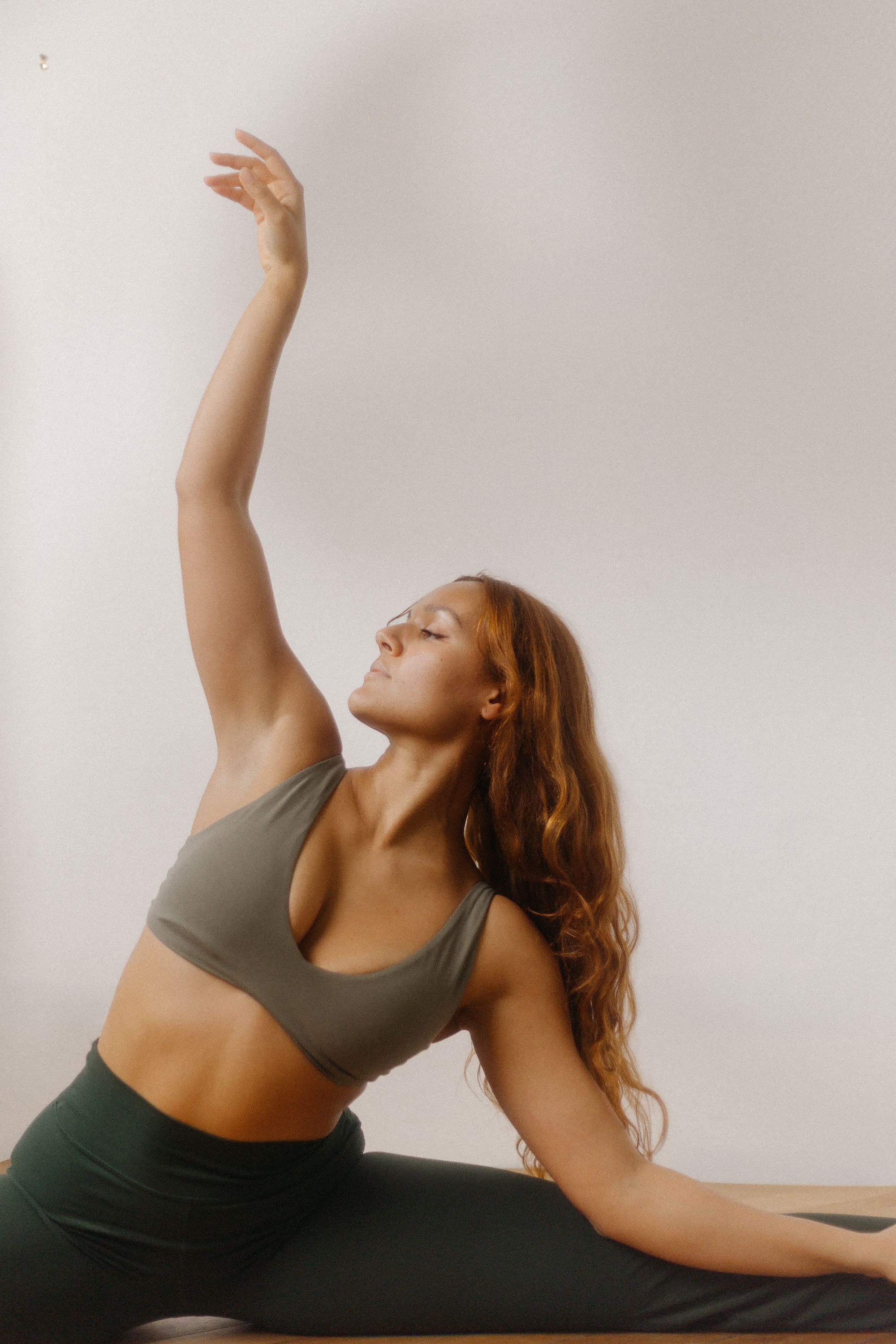 Woman with long red hair in green workout clothes stretching with one arm raised against a plain background.