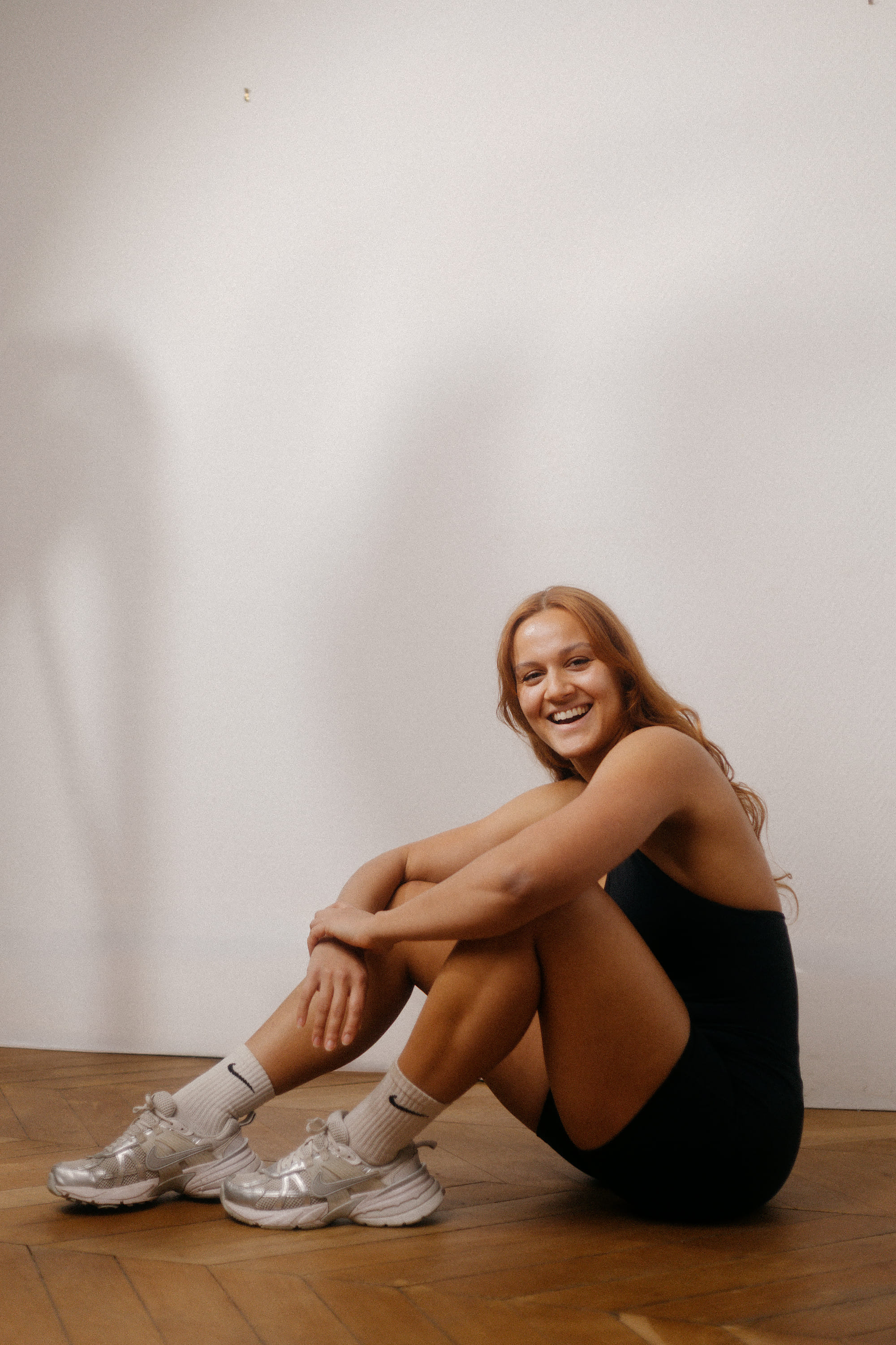 Smiling woman in black athletic outfit sitting cross-legged on wooden floor.