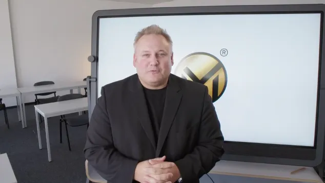 Im Bild ist Markus Mensch in einem schwarzen Anzug zu sehen, der vor einem großen Bildschirm steht, auf dem das Logo von Markus Mensch als goldfarbenes Symbol zu erkennen ist. Er hat seine Hände vor sich gefaltet und blickt direkt in die Kamera, was den Eindruck von Professionalität und Zuversicht vermittelt. Der Raum im Hintergrund scheint ein Büro oder Seminarraum zu sein, ausgestattet mit Tischen und Stühlen. 