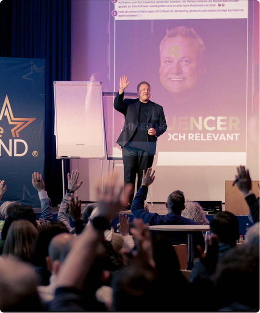 A speaker in a dark blazer raises his hand while addressing an audience with raised hands in a conference room.
