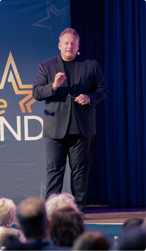 Man in a black suit speaking on stage in front of an audience, with a backdrop displaying a star and partial text.