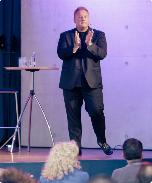 Man in black suit clapping on stage during a presentation with audience in foreground.