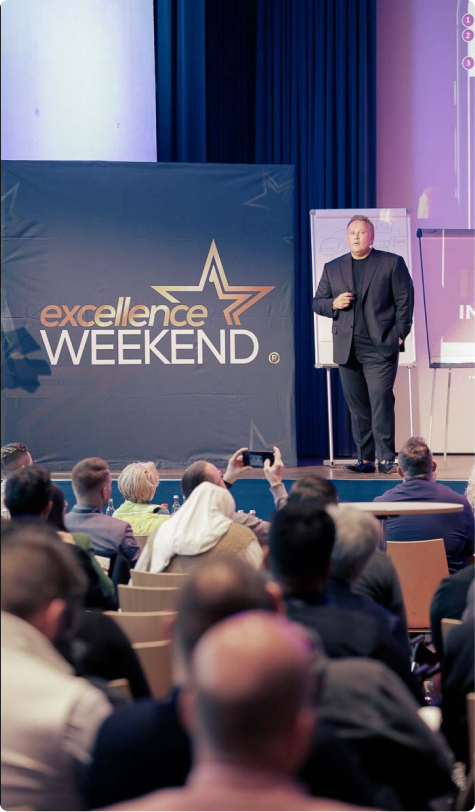 Speaker in black suit presenting on stage at an Excellence Weekend event with an audience watching.