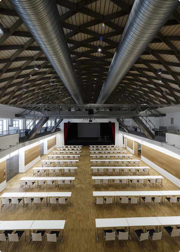 Large auditorium with rows of white tables and chairs facing a stage with red curtains under an exposed wooden beam ceiling and large ventilation ducts.