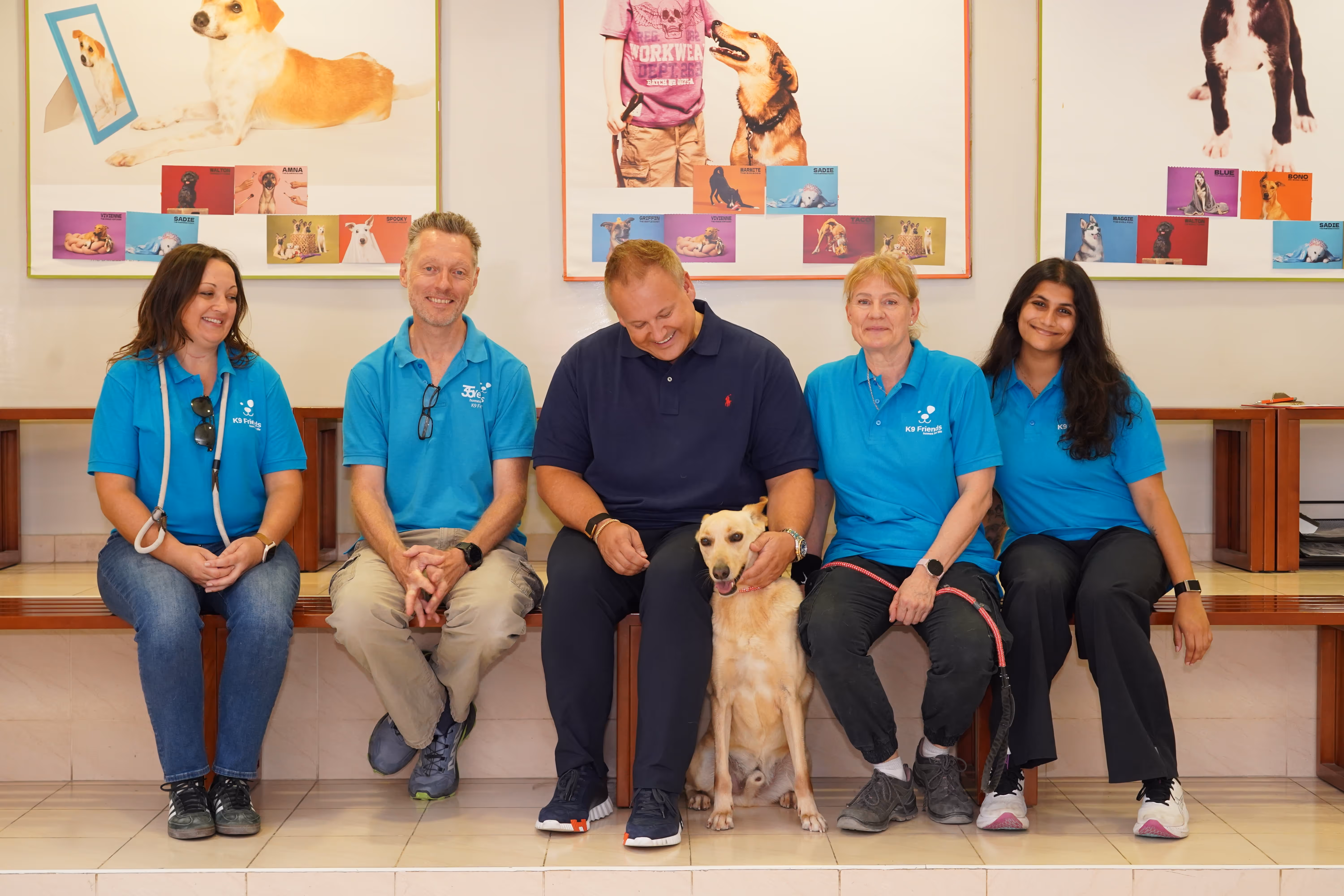 Fünf Personen sitzen mit einem Hund auf einer Bank, vier tragen blaue „K9 Friends“-T-Shirts; Markus Mensch sitzt in der Mitte und streichelt den Hund.
