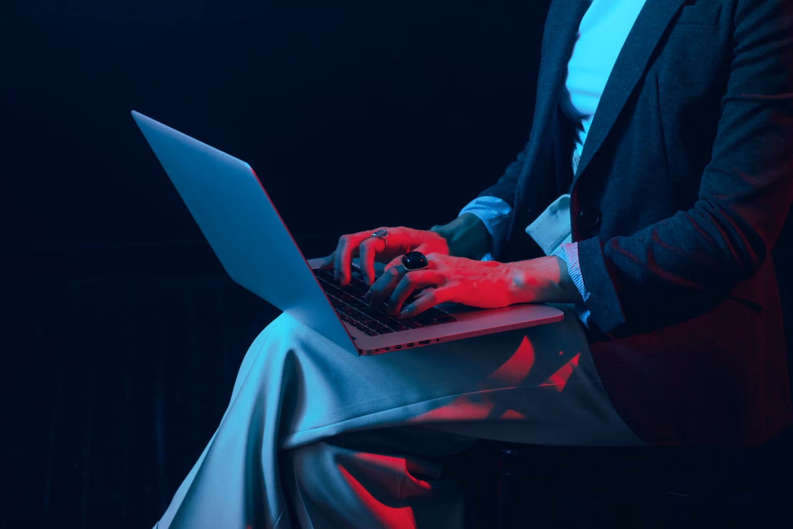 Close-up of hands typing on a laptop balanced on a lap, lit in dramatic blue and red tones, evoking working late at night on