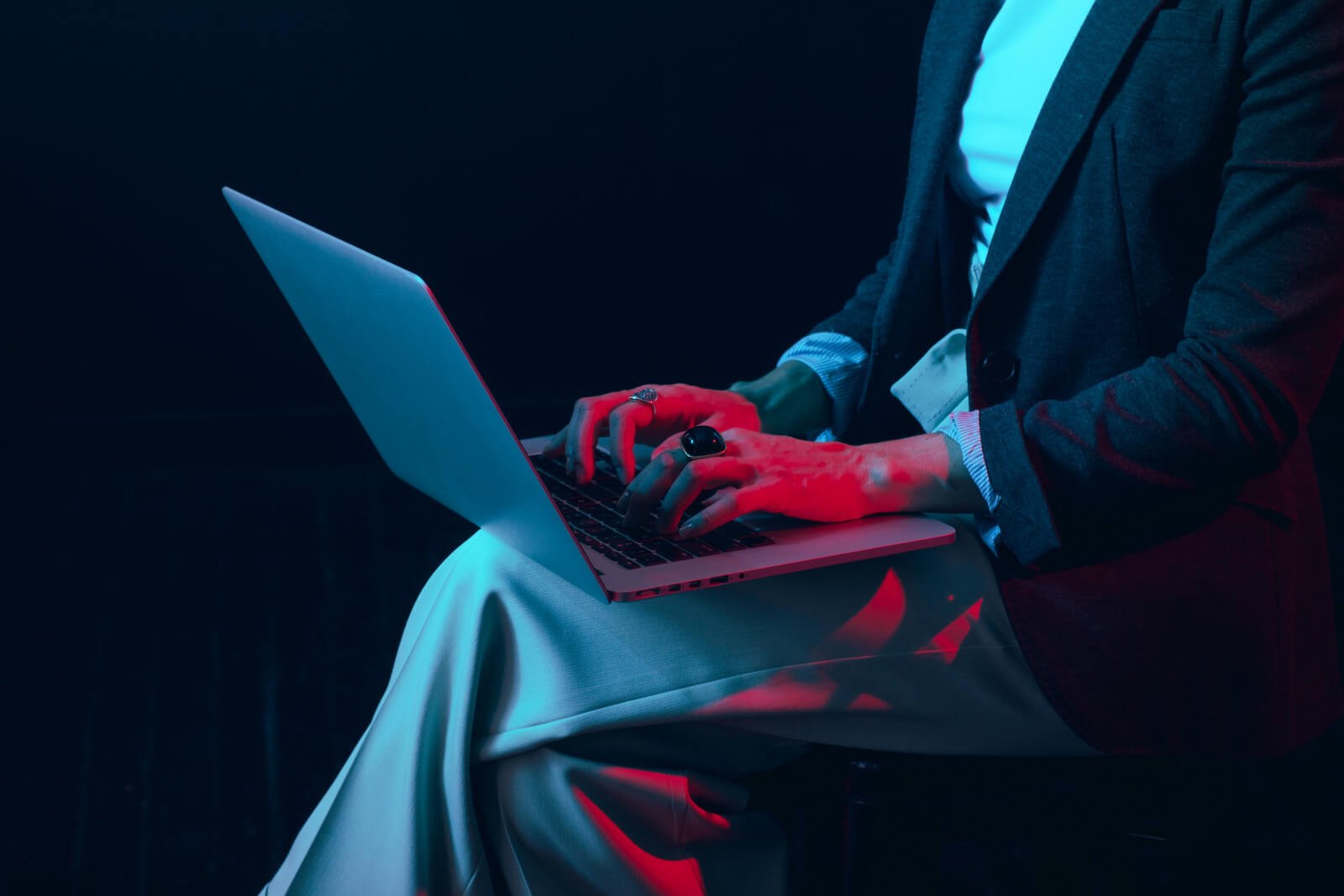 Close-up of hands typing on a laptop balanced on a lap, lit in dramatic blue and red tones, evoking working late at night on