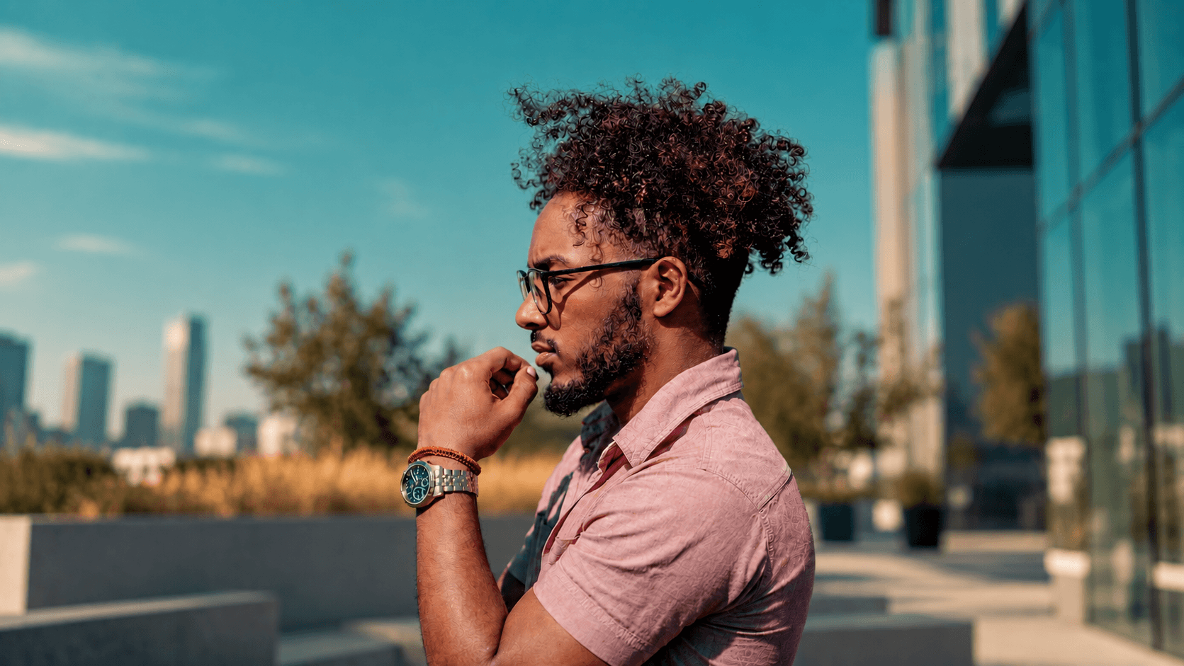 Man in glasses standing outside an office building, hand to his chin, looking thoughtfully into the distance.