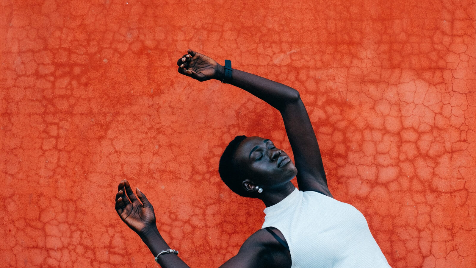 Woman in a white top with her arms raised and eyes closed, photographed against a cracked orange-red wall.