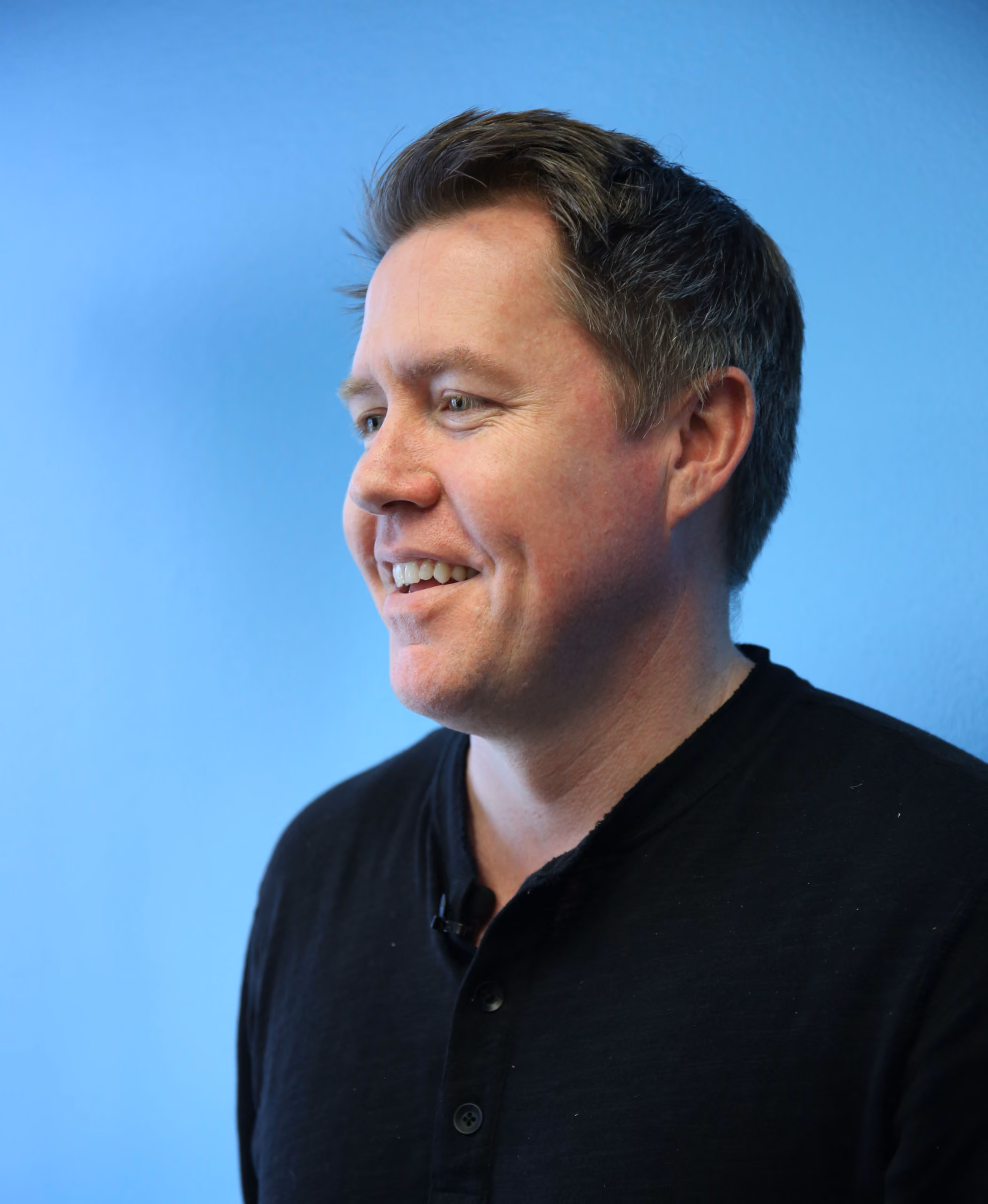 Smiling man with short brown hair wearing a black shirt, against a blue background.