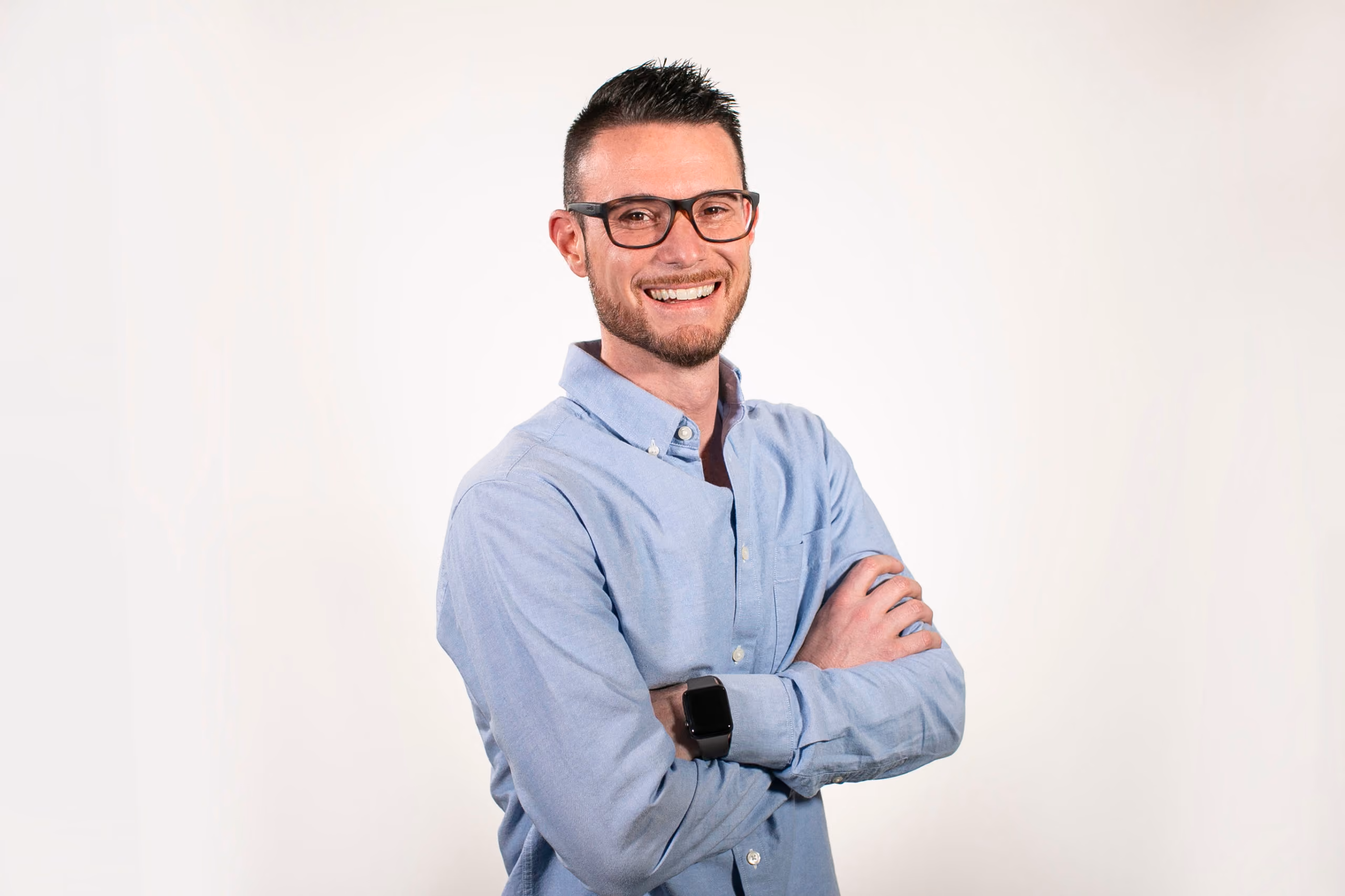 Smiling man with short dark hair and glasses wearing a light blue shirt, arms crossed.