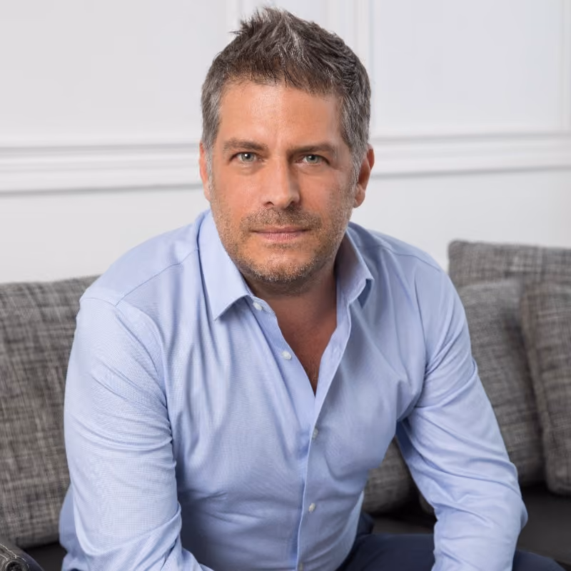 Man with short gray hair and stubble wearing a light blue shirt sitting on a gray couch.