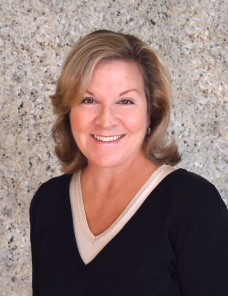 Smiling woman with blonde hair wearing a black sweater over a beige top against a speckled gray wall.