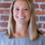 Smiling woman with shoulder-length blonde hair in light blue top against brick wall background.