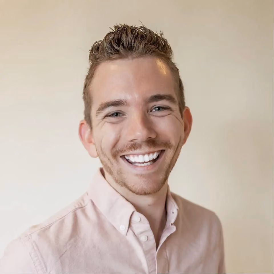 Smiling young man with short hair wearing a light pink button-up shirt against a neutral background.