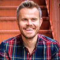 Smiling man with short blonde hair wearing a plaid shirt sitting on red stairs.