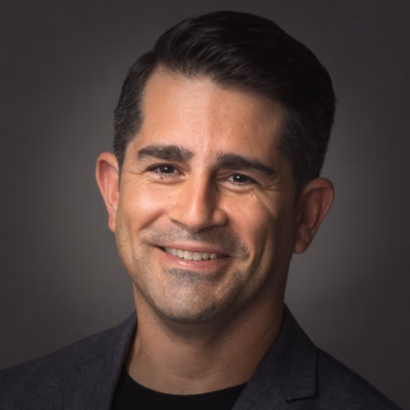 Smiling man with dark hair wearing a dark blazer over a black shirt against a gray background.