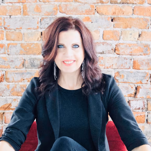 Smiling woman with wavy dark hair and blue eyes wearing a black blazer sitting in front of a brick wall.