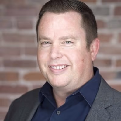 Smiling man with short hair wearing a navy shirt and gray blazer against a blurred brick wall background.