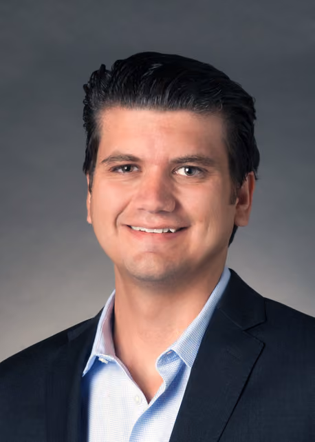 Smiling man with dark hair wearing a black blazer and light blue shirt against gray background.