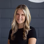 Smiling woman with long blonde hair wearing a black shirt in front of a dark gray wall.