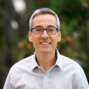 Smiling man with glasses and short gray hair wearing a light checkered shirt outdoors.