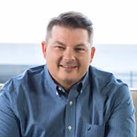 Smiling man with short brown hair wearing a blue checkered shirt against a bright background.