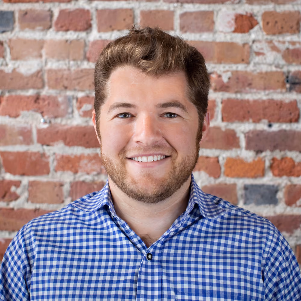 Smiling man with short hair and beard wearing a blue checkered shirt in front of a brick wall.