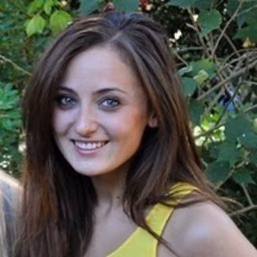 Smiling woman with long brown hair wearing a yellow top standing outdoors near greenery.