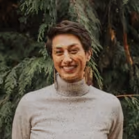 Smiling person with short brown hair and wooden earrings standing outdoors with green foliage backdrop.