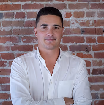 Man in a white button-up shirt standing with arms crossed in front of a red brick wall.