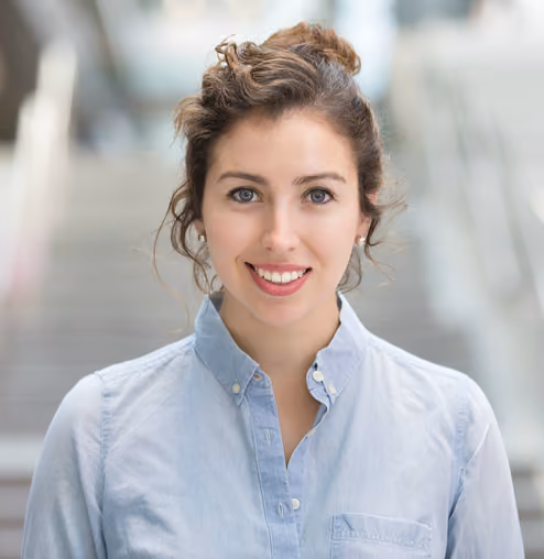 Smiling young woman with light blue button-up shirt and hair tied back in a blurred indoor setting.