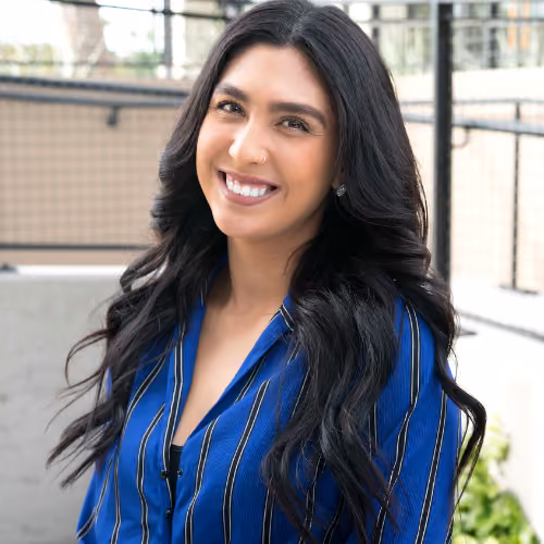 Smiling woman with long black hair wearing a blue striped shirt, standing outdoors in daytime.