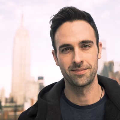 Smiling man with dark hair wearing a black jacket with a city skyline in soft focus behind him.